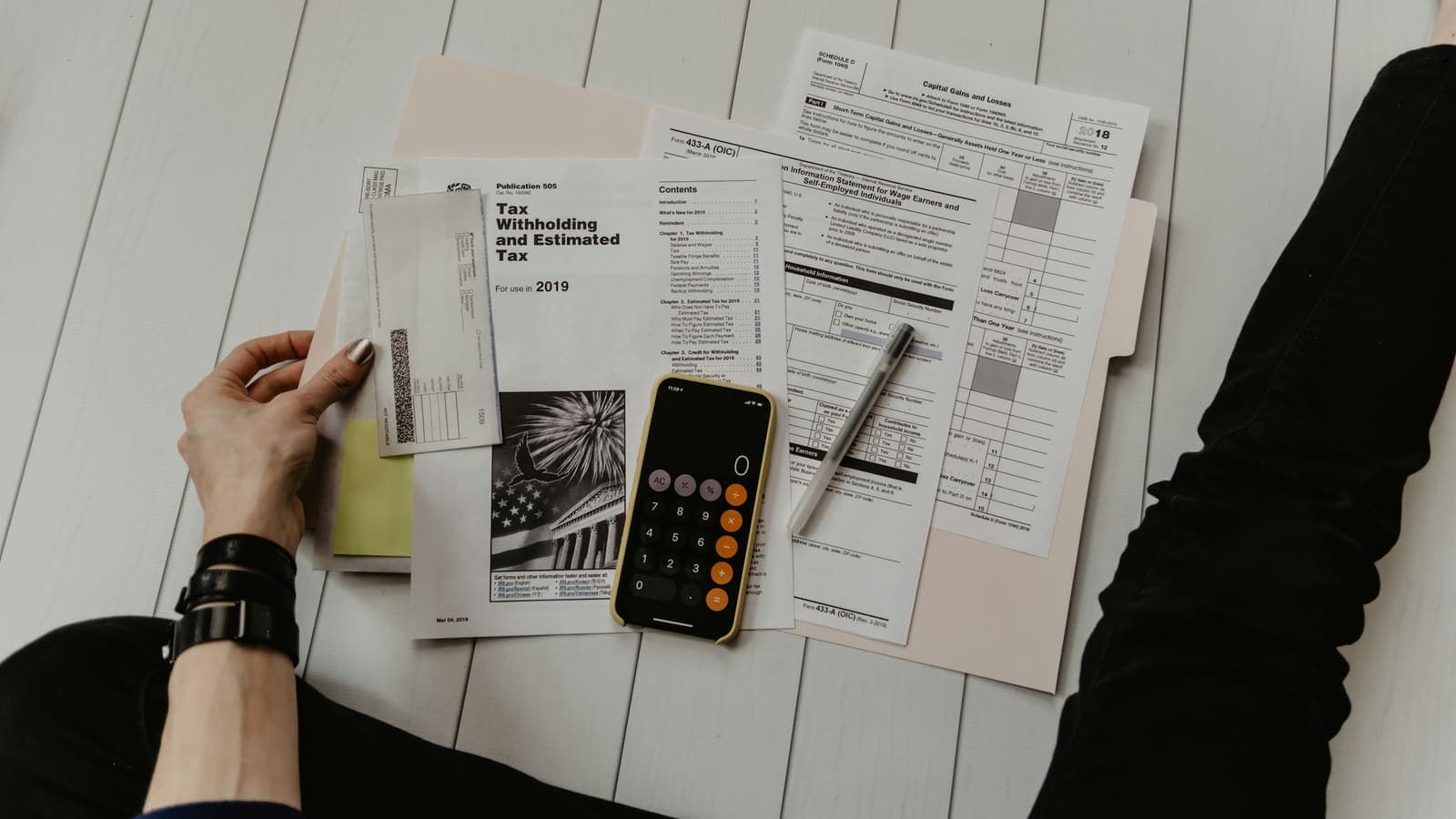 A person sorting IRS Publication 505 and a Schedule D form across a table, with a calculator open on a phone beside the papers.