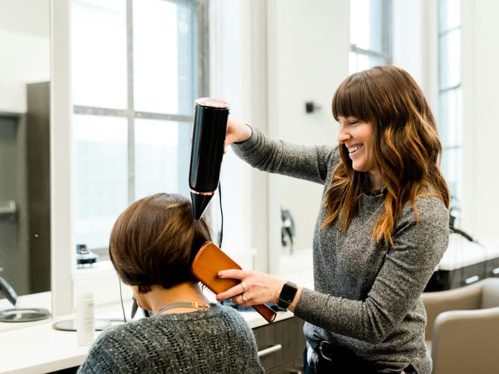 A hair stylist blow-drying and brushing out a client's hair at a salon chair.
